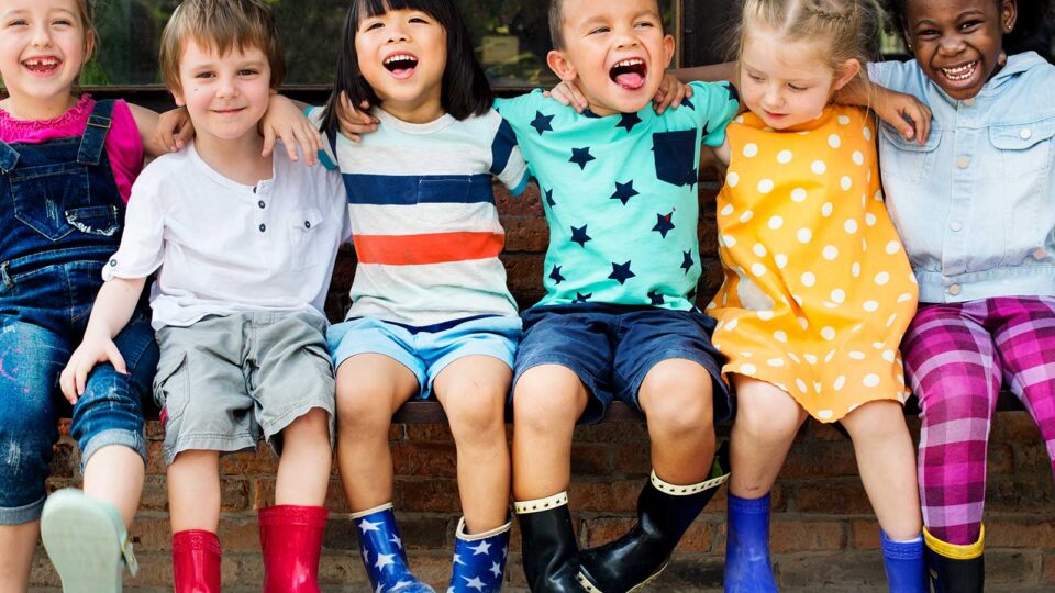 A diverse group of six children sits closely together, smiling and wearing colorful outfits, including bright boots and playful patterns.