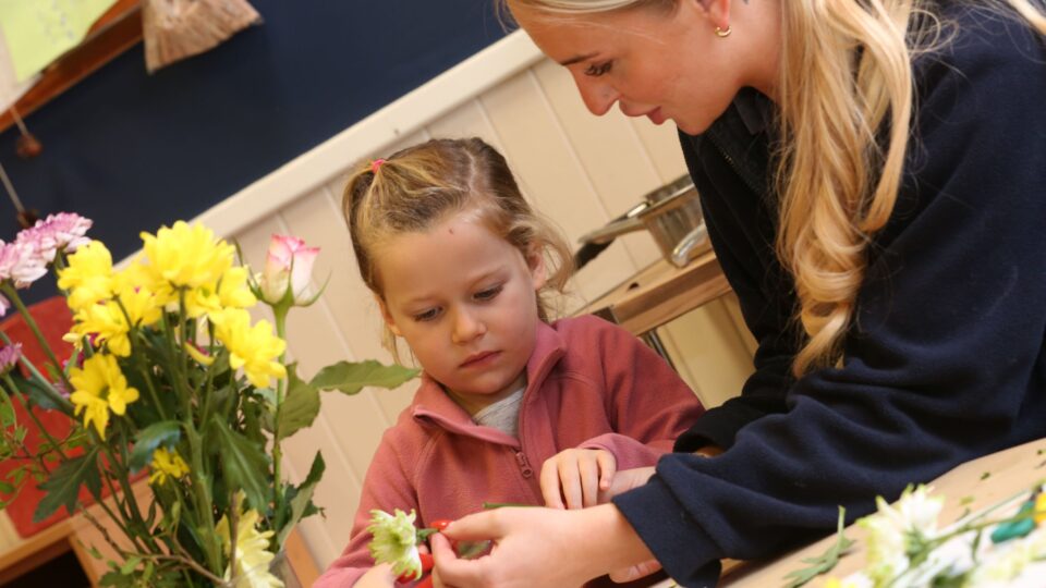 A young girl and an educator craft together, surrounded by colorful flowers, fostering creativity and hands-on learning.