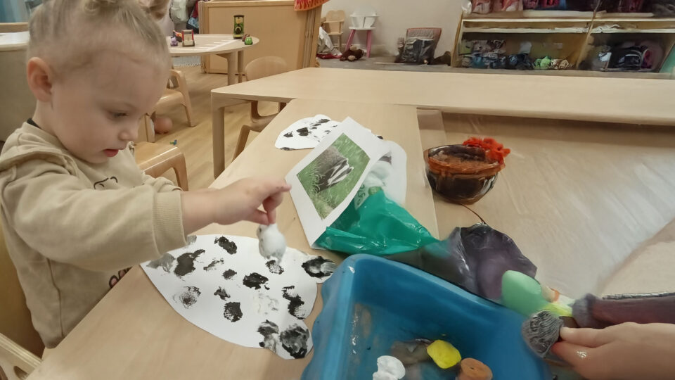 A child creates art with black and white paint on paper, surrounded by colorful materials and a picture of a badger in a nursery setting.