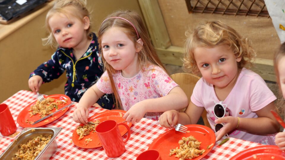 Three young children at a West Sussex nursery enjoy a nutritious pasta meal, sitting together at a red-checked table.