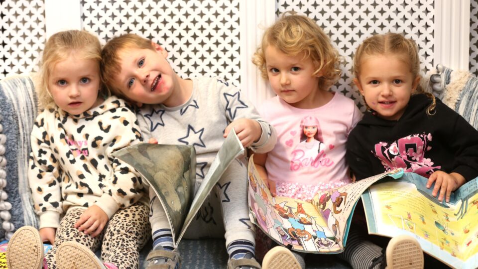 Four children sit together on a cozy couch, each holding a colorful book. They wear playful, varied outfits and exhibit joyful expressions.